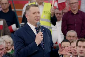 Przemysław Czarnek podczas wystąpienia publicznego w Krakowie, w tle grafika symbolizująca odnawialne źródła energii i dom z panelami fotowoltaicznymi