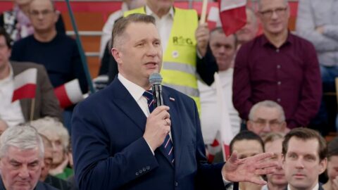 Przemysław Czarnek podczas wystąpienia publicznego w Krakowie, w tle grafika symbolizująca odnawialne źródła energii i dom z panelami fotowoltaicznymi